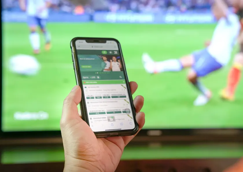 Phone showing a betting app beside a football match on TV, illustrating sports betting in Malaysia